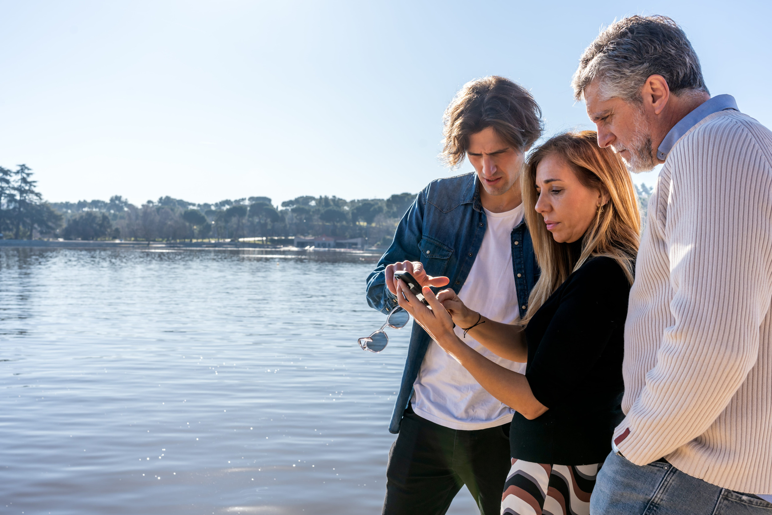 Step 1 sell Selling a Home on Lake Lanier: Side view of mature mother with cellphone showing photos while young son pointing at screen and husband standing with hand in pocket near river