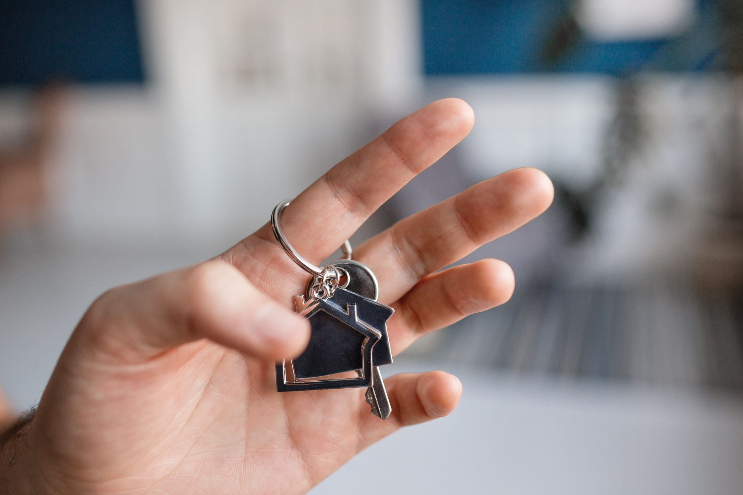 Buying a Home on Lake Lanier: Men hand holding key with house shaped keychain. Modern light lobby interior. Mortgage concept. Real estate, moving home or renting property.